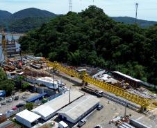 Imagem aérea de das obras da ponte de guaratuba em andamento em uma região costeira, com maquinário e áreas de construção cercadas por vegetação exuberante e montanhas ao fundo.