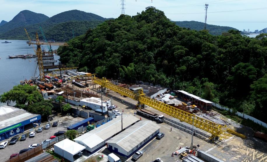 Imagem aérea de das obras da ponte de guaratuba em andamento em uma região costeira, com maquinário e áreas de construção cercadas por vegetação exuberante e montanhas ao fundo.